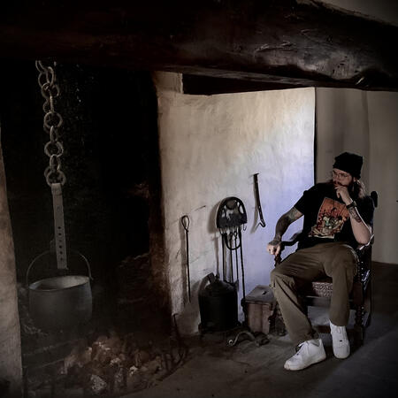 Rev. Árón at the hearth. A modern Irish practitioner of folk magic sits beside a hearth in a traditional cottage, surrounded by tools of spiritwork and homecraft. A quiet moment of reflection and connection to ancestral ways.